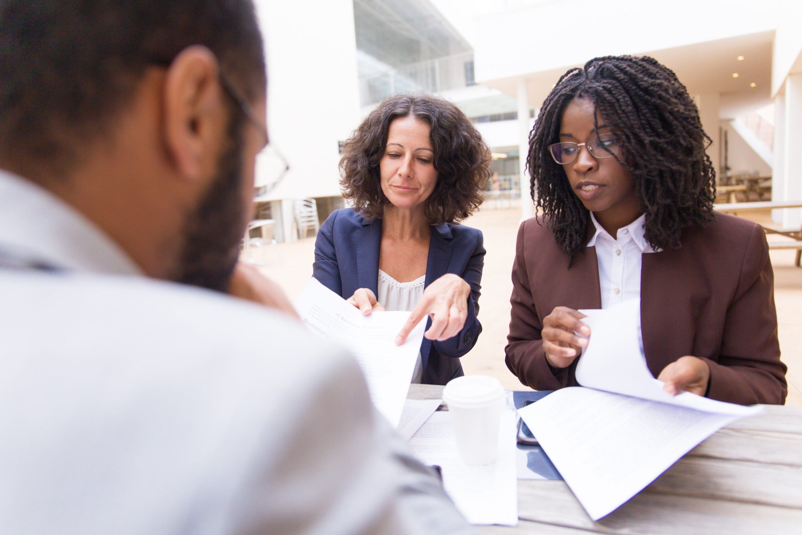 Mix raced partners discussing agreement terms in outdoor cafe. Business women sitting at table outside, showing documents to male colleague. Meeting or negotiation concept